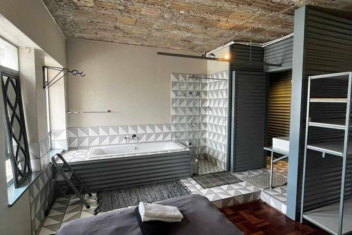 Unique bedroom featuring an open-plan ensuite with a built-in bathtub, glass shower, exposed brick ceiling, and industrial-style corrugated metal accents.