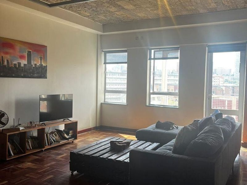 Spacious living room featuring industrial-style exposed brick ceiling, rich parquet flooring, and large windows providing ample natural light and urban views.