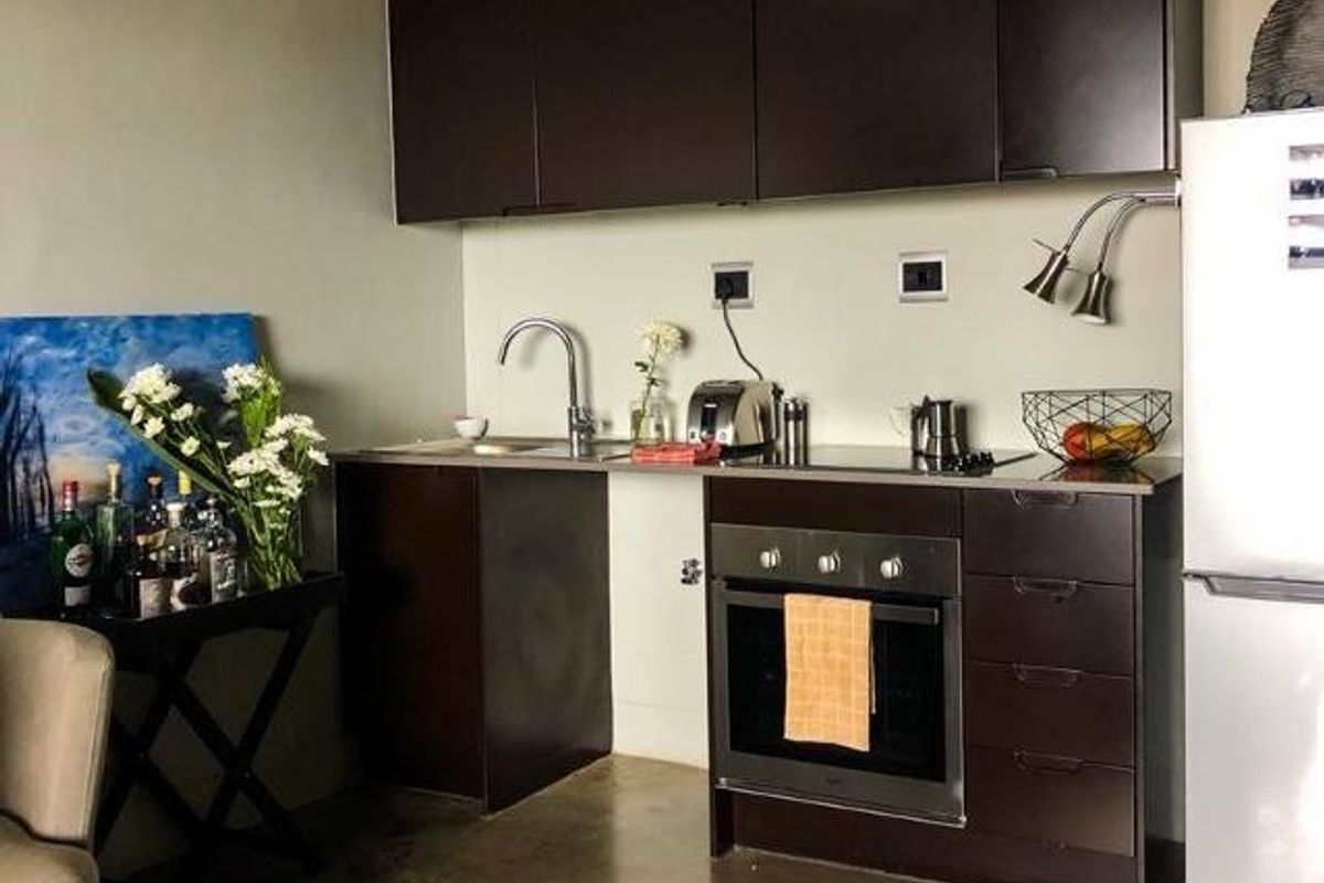 Compact industrial-style kitchen featuring dark cabinetry, stainless steel sink, built-in oven, electric hob, and polished concrete flooring with exposed ceiling ducting.