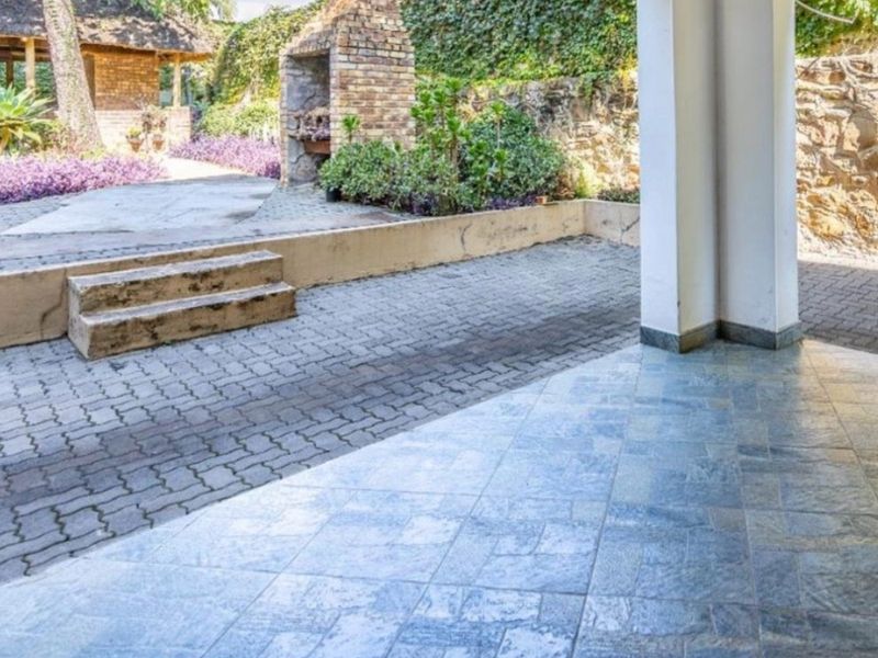 Covered patio area featuring tiled flooring, a brick built-in braai, and direct access to a paved garden space with stone retaining walls.