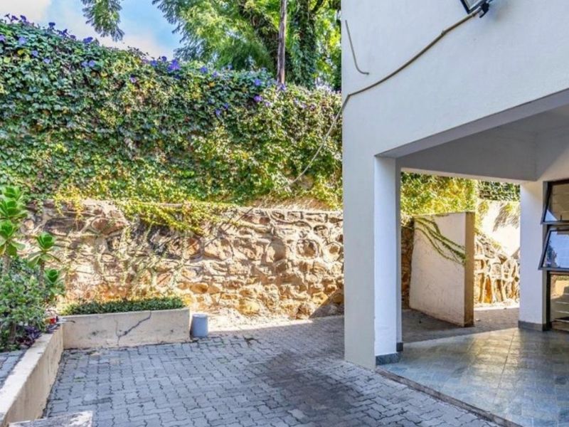 A covered patio area featuring tiled flooring, adjacent to a stone retaining wall with established climbing plants and a paved outdoor space.
