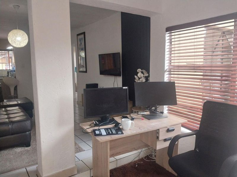 Open plan living area featuring tiled flooring, decorative wall paneling, and large windows with venetian blinds, providing a functional space for living and work.