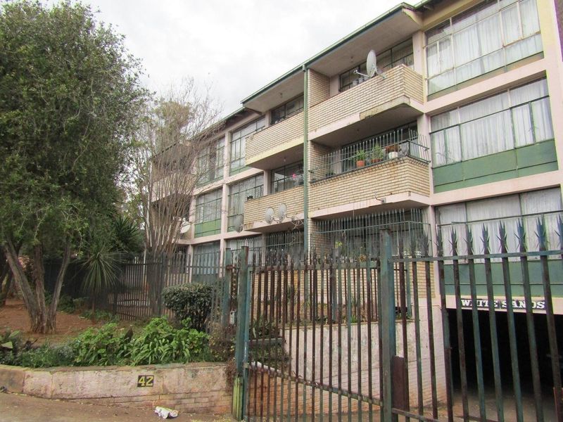 Exterior view of a multi-story apartment building featuring brick facade, private balconies, and secure perimeter fencing with an underground parking entrance.