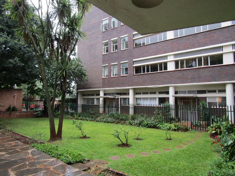 A view of the apartment building exterior featuring a brick facade, metal security fencing, and a well-maintained communal garden area with a stone walkway.