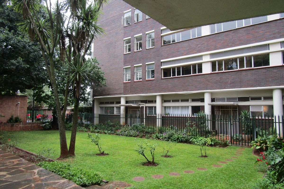A view of the apartment building exterior featuring a brick facade, metal security fencing, and a well-maintained communal garden area with a stone walkway.