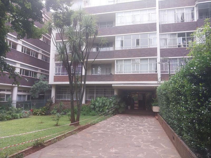 Front view of a multi-story brick apartment building featuring a paved driveway, landscaped garden area, and balconies with security bars.