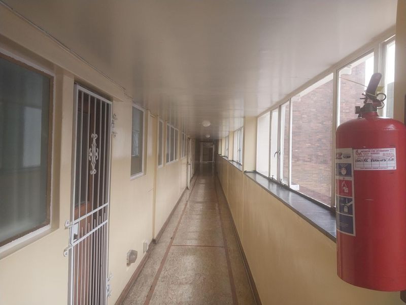 A long, enclosed apartment building hallway featuring tiled flooring, security gates, and large windows providing natural light.