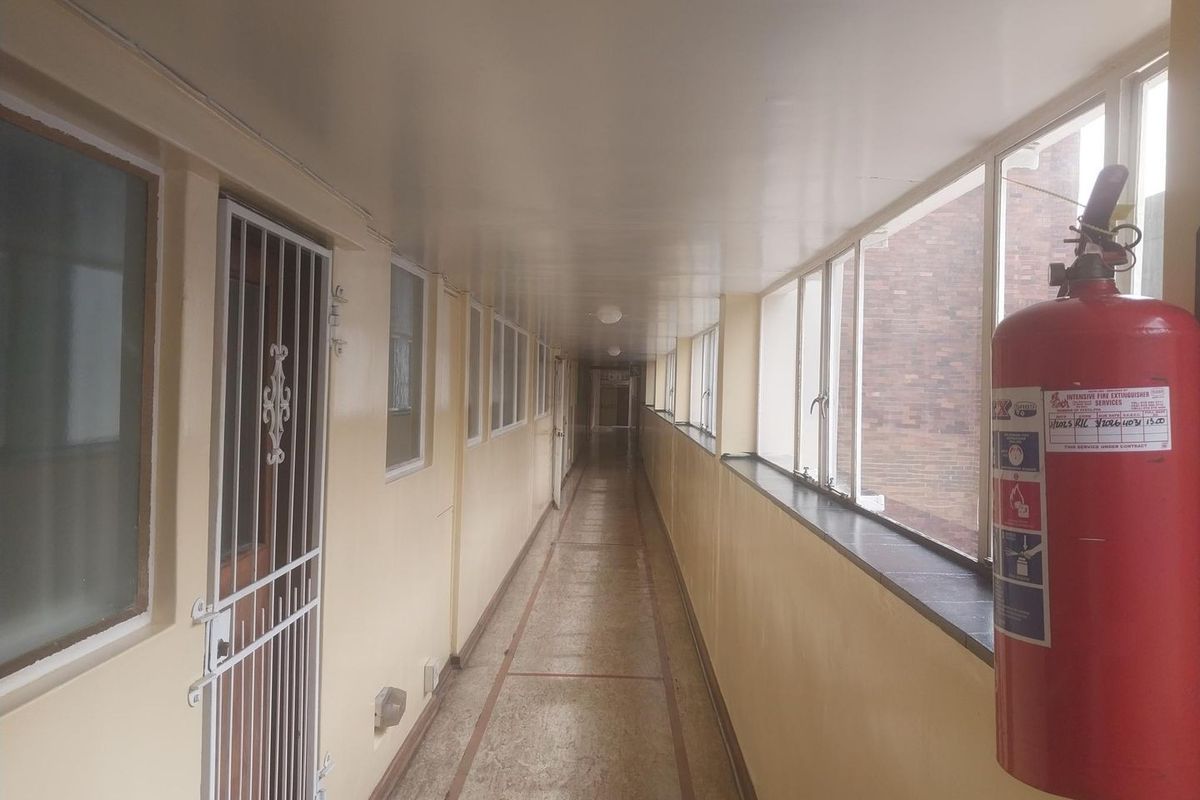 A long, enclosed apartment building hallway featuring tiled flooring, security gates, and large windows providing natural light.