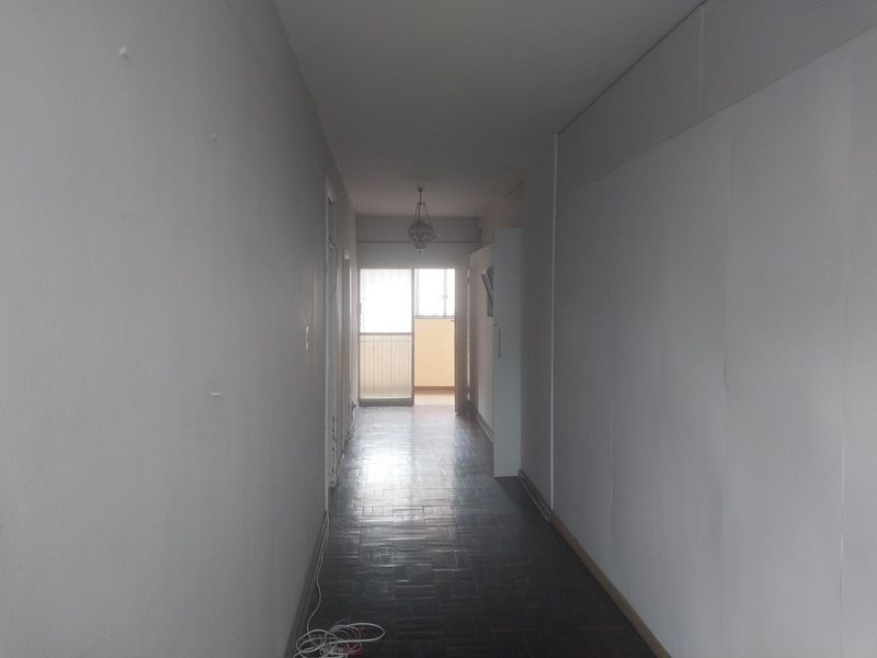 A vacant hallway featuring dark parquet flooring and plain walls, leading towards a bright doorway at the end of the corridor.
