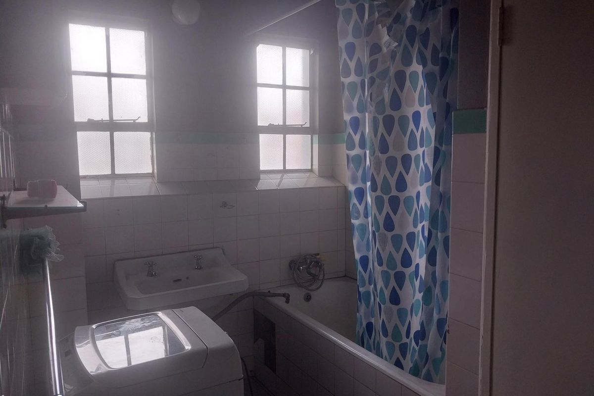 A dated bathroom featuring a wall-mounted basin, bathtub with shower curtain, and tiled walls, requiring modernization and repair of the bath panel.