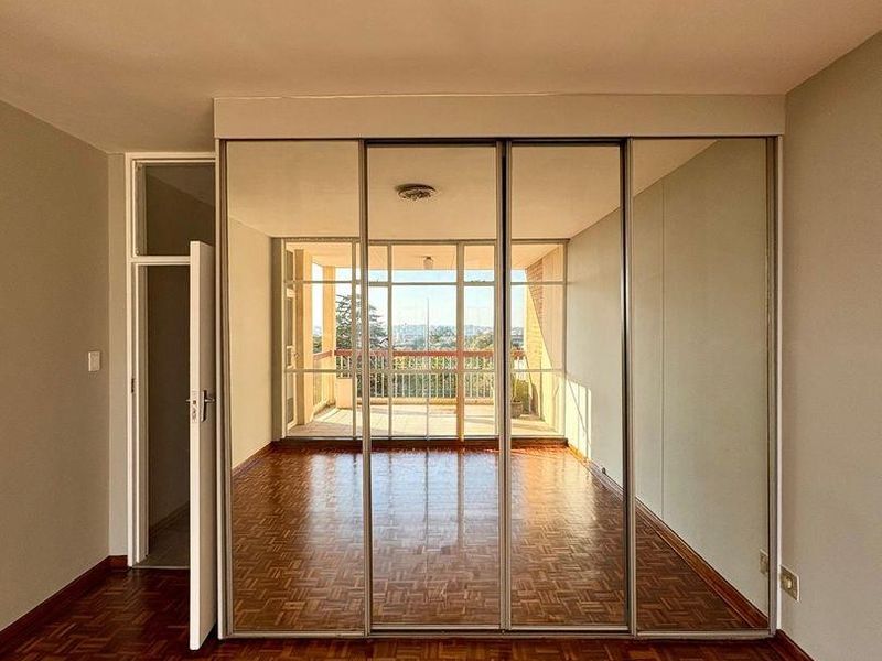 A bright, empty bedroom featuring classic parquet flooring and large floor-to-ceiling mirrored sliding wardrobe doors, with a view through to an adjacent balcony.
