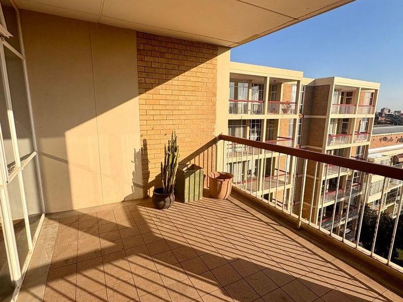 A covered balcony featuring tiled flooring, a metal safety railing, and brick wall accents, offering an open outdoor space with views of the apartment complex.