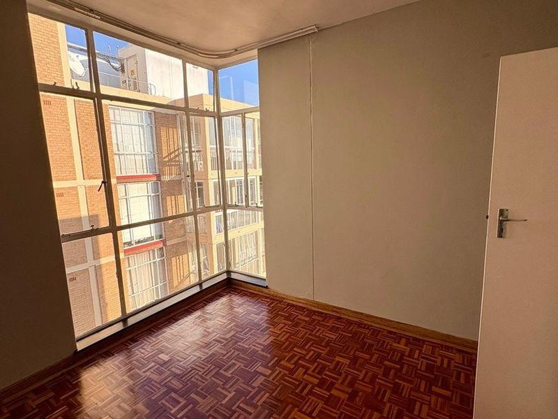 An empty bedroom featuring classic parquet flooring and a large corner window providing natural light and urban views.