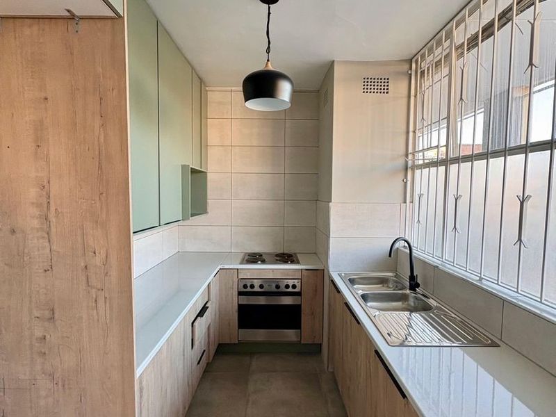 Modern galley-style kitchen featuring light wood-grain cabinetry, stone countertops, integrated stainless steel oven and hob, and a double sink with a matte black faucet.