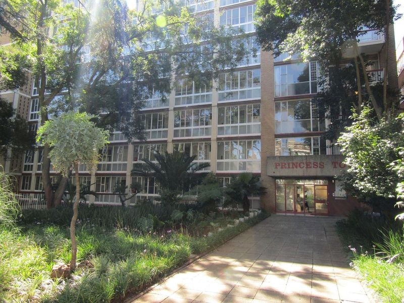 Front exterior view of Princess Towers apartment building featuring a paved entrance walkway, landscaped garden, and multi-story brick facade with large windows.