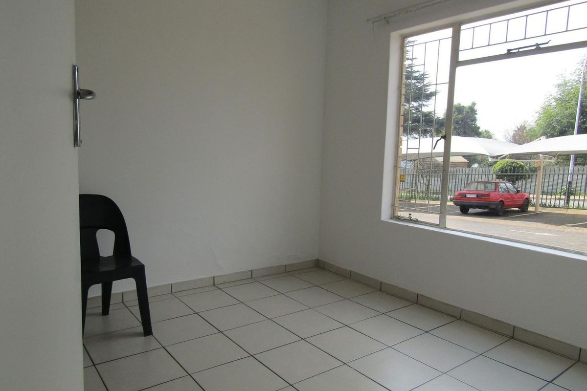 A bright, empty living area featuring neutral tiled flooring and a large window with security bars, offering a view of the parking area.