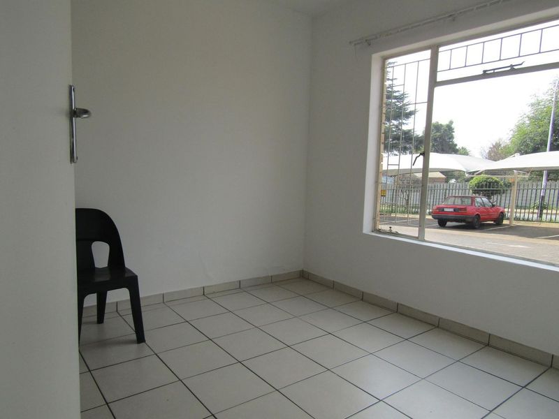 A bright, empty living area featuring neutral tiled flooring and a large window with security bars, offering a view of the parking area.