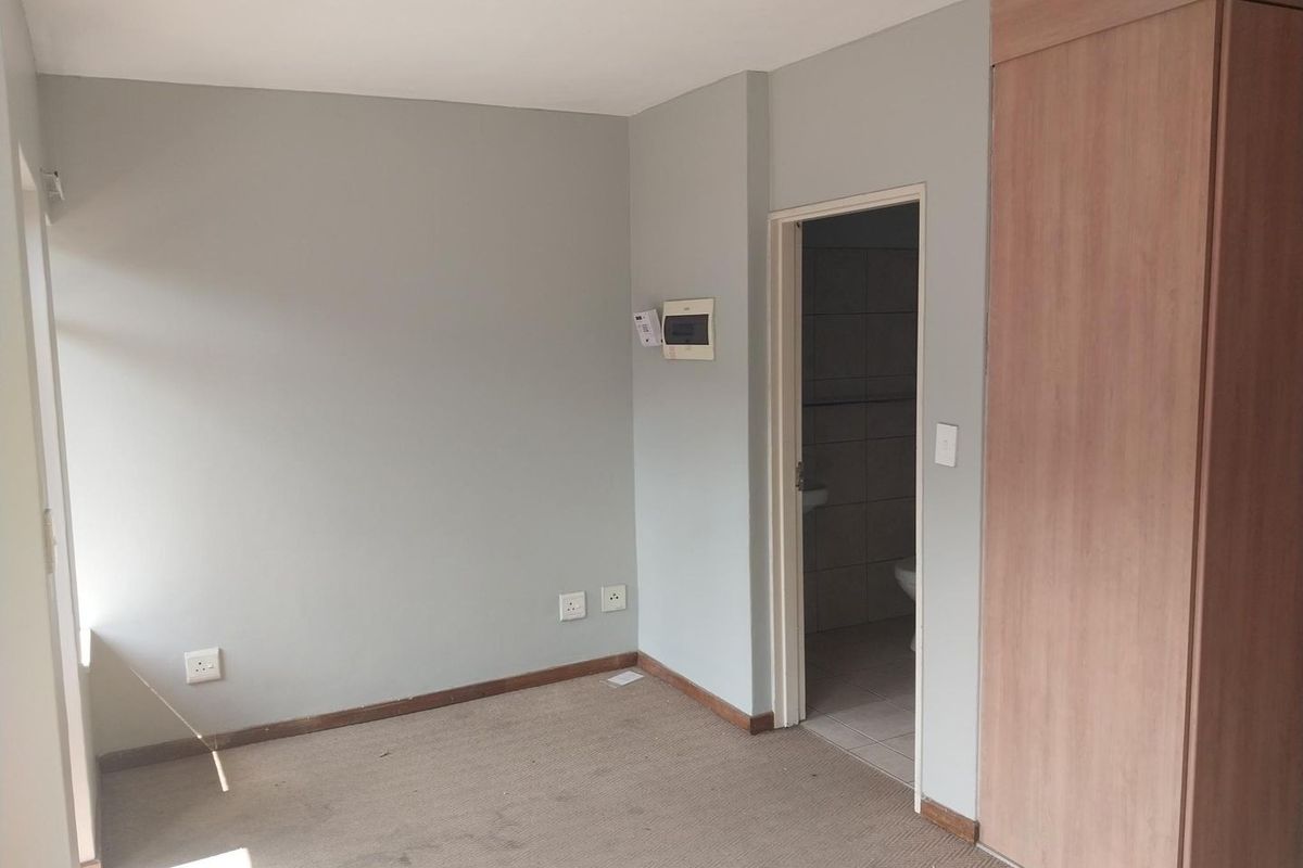 An empty bedroom featuring neutral walls, built-in wooden cupboards, and carpeted flooring, with an adjacent bathroom visible through the doorway.