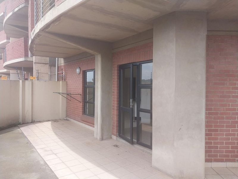 A covered ground floor patio featuring exposed brick walls, concrete pillars, tiled flooring, and an aluminium framed glass entrance door.