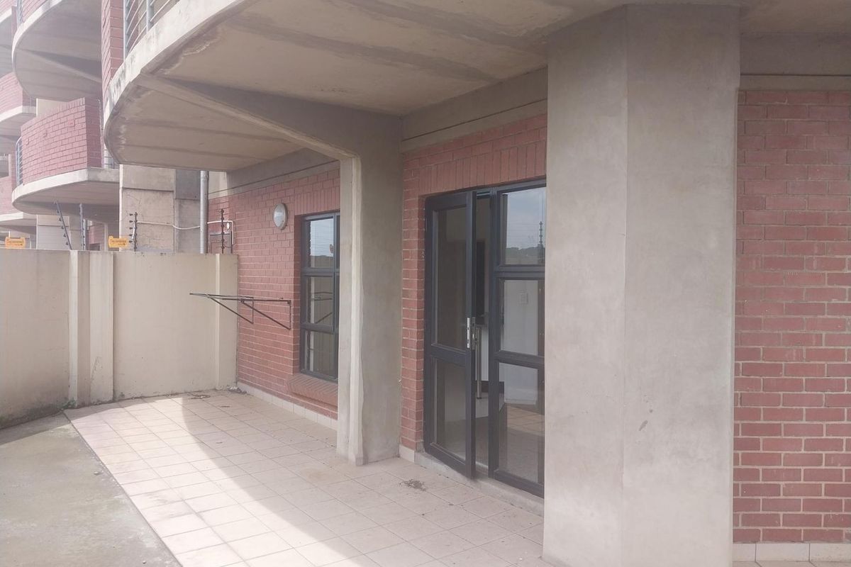 A covered ground floor patio featuring exposed brick walls, concrete pillars, tiled flooring, and an aluminium framed glass entrance door.