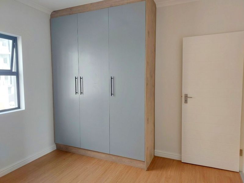 A clean, empty bedroom featuring modern built-in wardrobes with a light wood and grey finish, laminate flooring, and a neutral color palette.