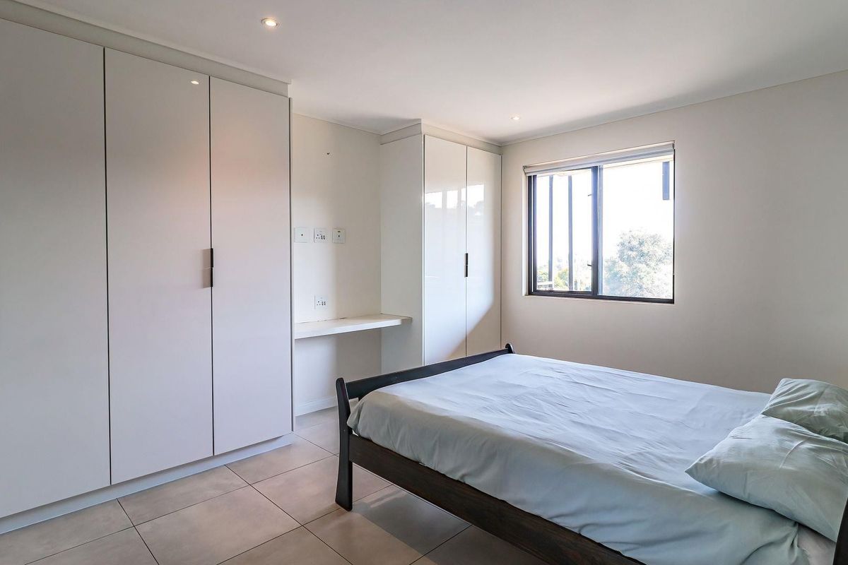 A bright bedroom featuring neutral tiled flooring, extensive built-in cabinetry with an integrated desk, and security bars on the window.