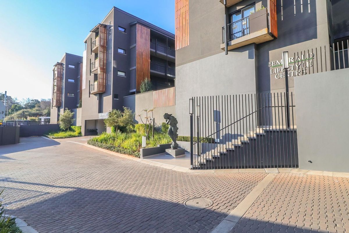 Exterior view of the modern apartment complex entrance, featuring paved driveway, secure metal fencing, and contemporary architectural facade details.