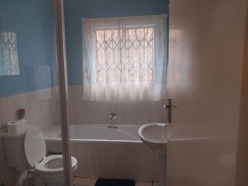 A functional bathroom featuring a bathtub, pedestal basin, and toilet, with tiled finishes and a window fitted with a security gate.