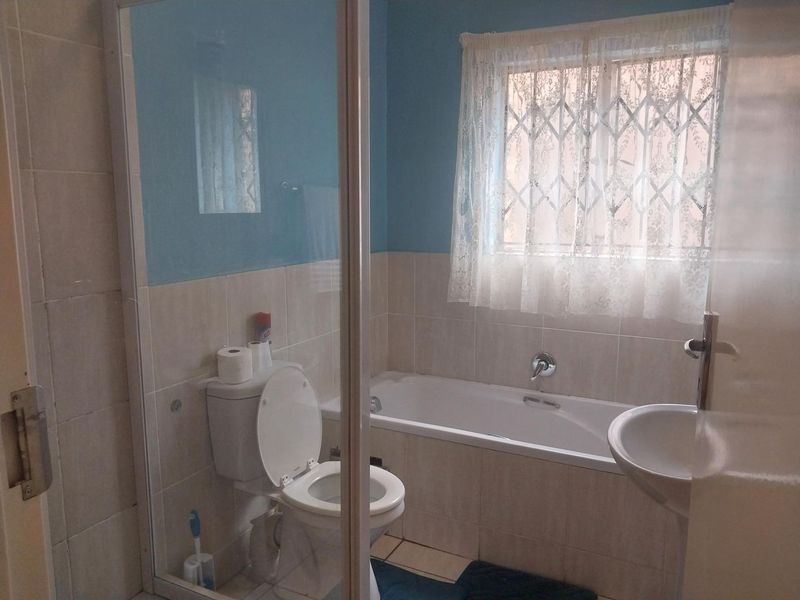 A functional bathroom featuring a bathtub, pedestal basin, toilet, and a shower enclosure with tiled finishes and a window with security bars.