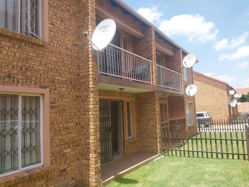 Exterior view of a face brick apartment building featuring a ground floor patio and an upper-level balcony with metal security railings and window bars.