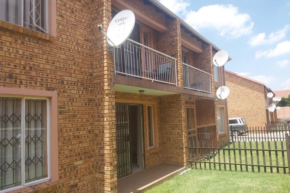 Exterior view of a face brick apartment building featuring a ground floor patio and an upper-level balcony with metal security railings and window bars.