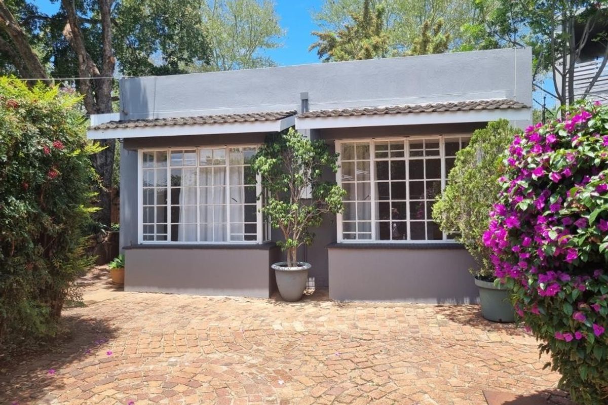 A well-maintained rear exterior view featuring a paved courtyard, rendered masonry walls, and classic multi-pane windows set against a lush garden backdrop.