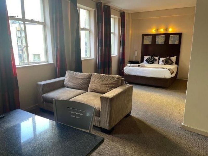 An open-plan apartment bedroom and living area featuring large windows, neutral wall finishes, and carpeted flooring.