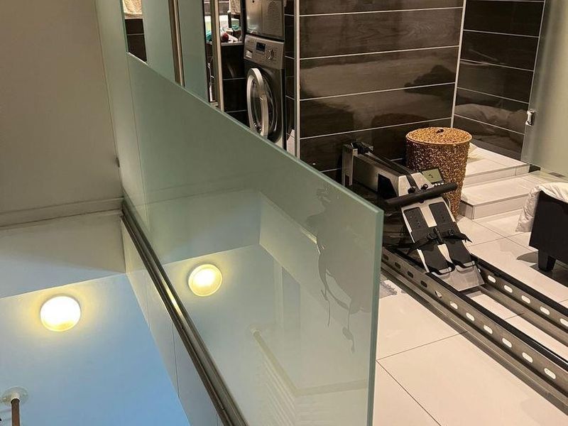 A modern laundry area featuring stacked appliances, sleek dark wall tiling, and a contemporary glass balustrade overlooking the staircase.