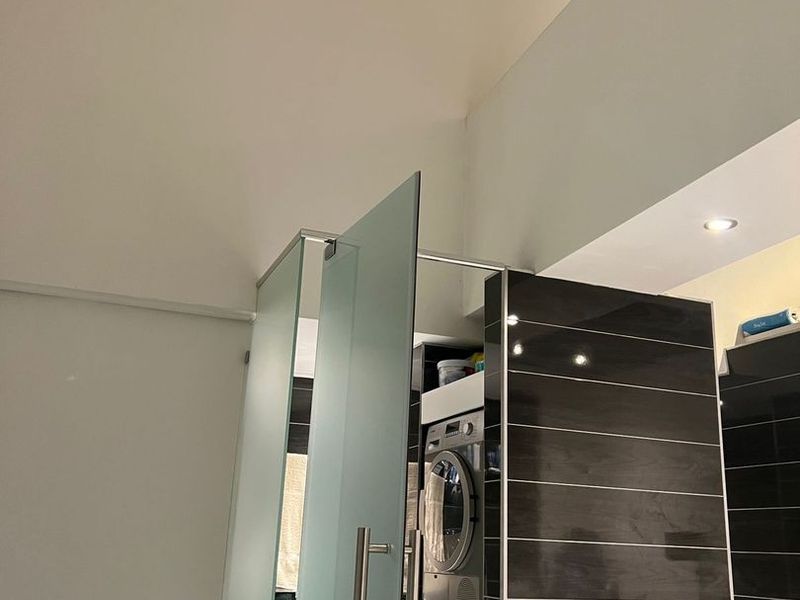 A modern laundry area featuring a stacked washer and dryer unit, sleek dark tiled wall, and a contemporary frosted glass privacy screen.