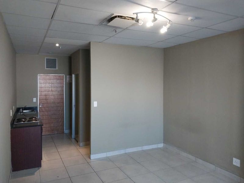 An empty open-plan living area featuring tiled floors, a small kitchenette with a hob and sink, and a drop ceiling with visible damage.