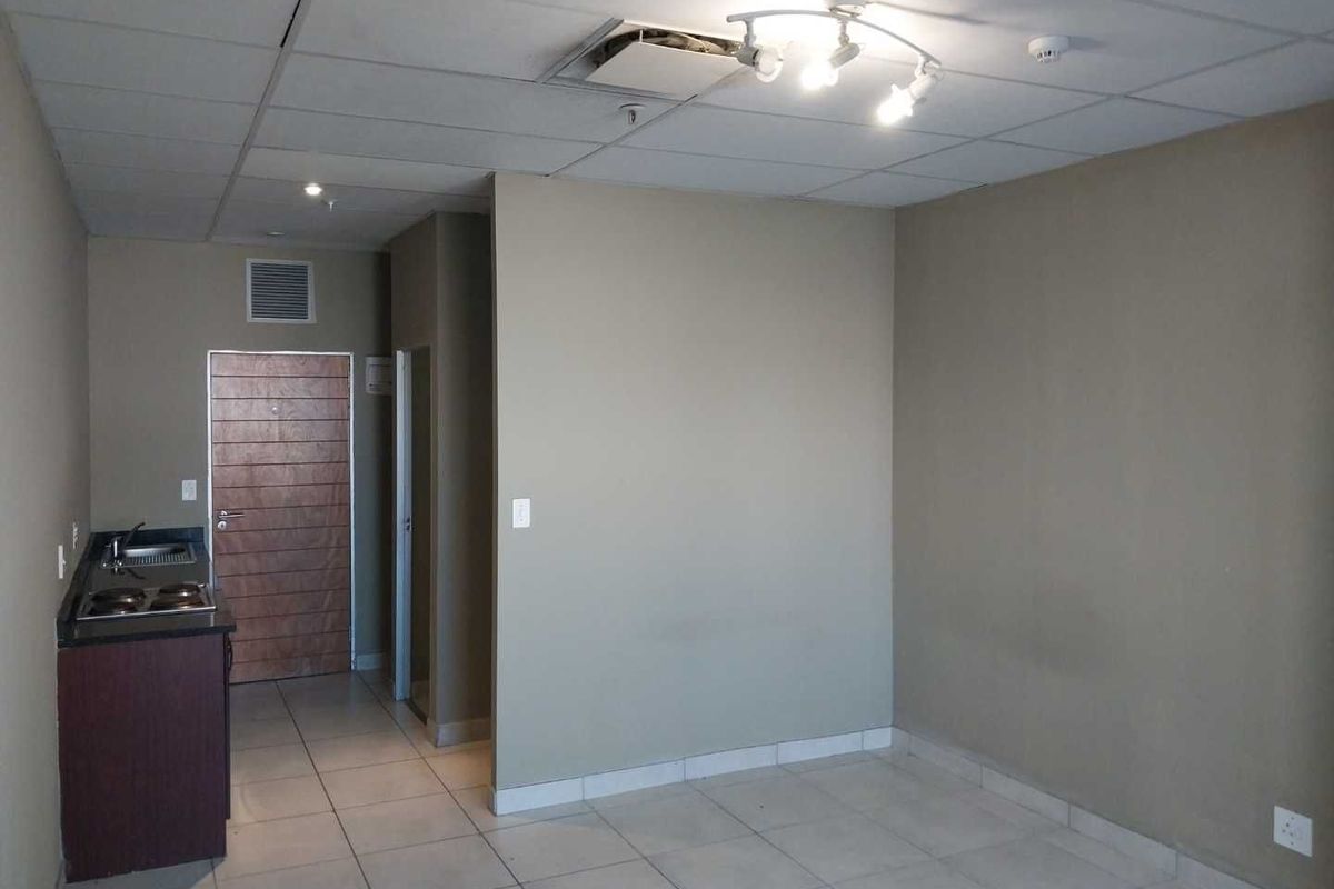 An empty open-plan living area featuring tiled floors, a small kitchenette with a hob and sink, and a drop ceiling with visible damage.