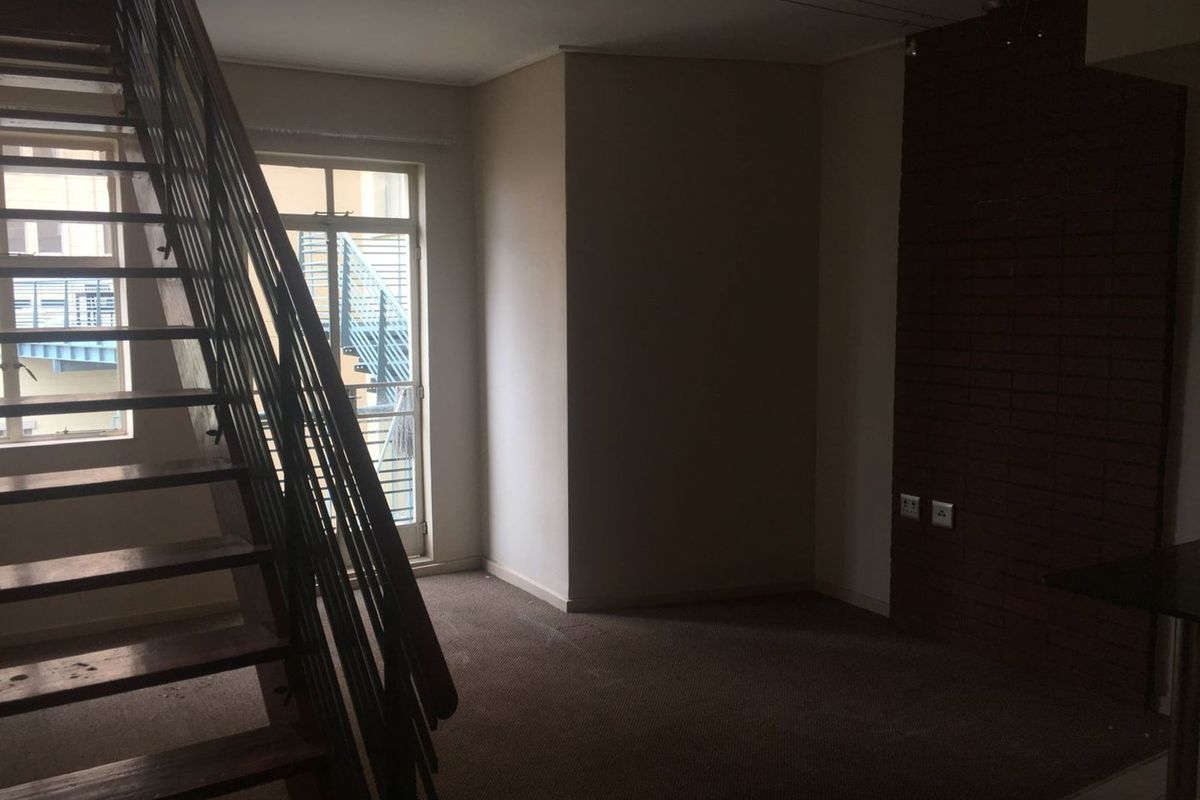 An empty living area featuring an exposed brick wall, carpeted flooring, and an internal staircase leading to the upper level.