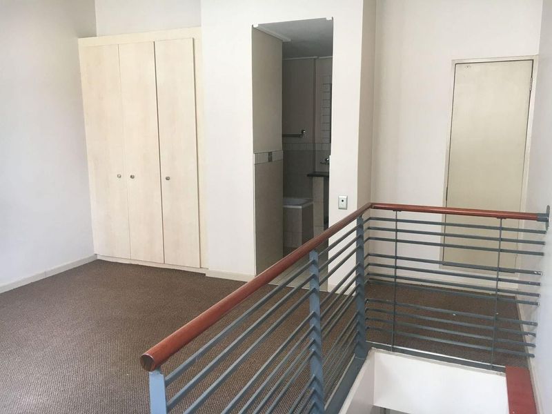An empty bedroom featuring built-in cupboards, carpeted flooring, and a metal balustrade overlooking the staircase, with an adjacent en-suite bathroom.