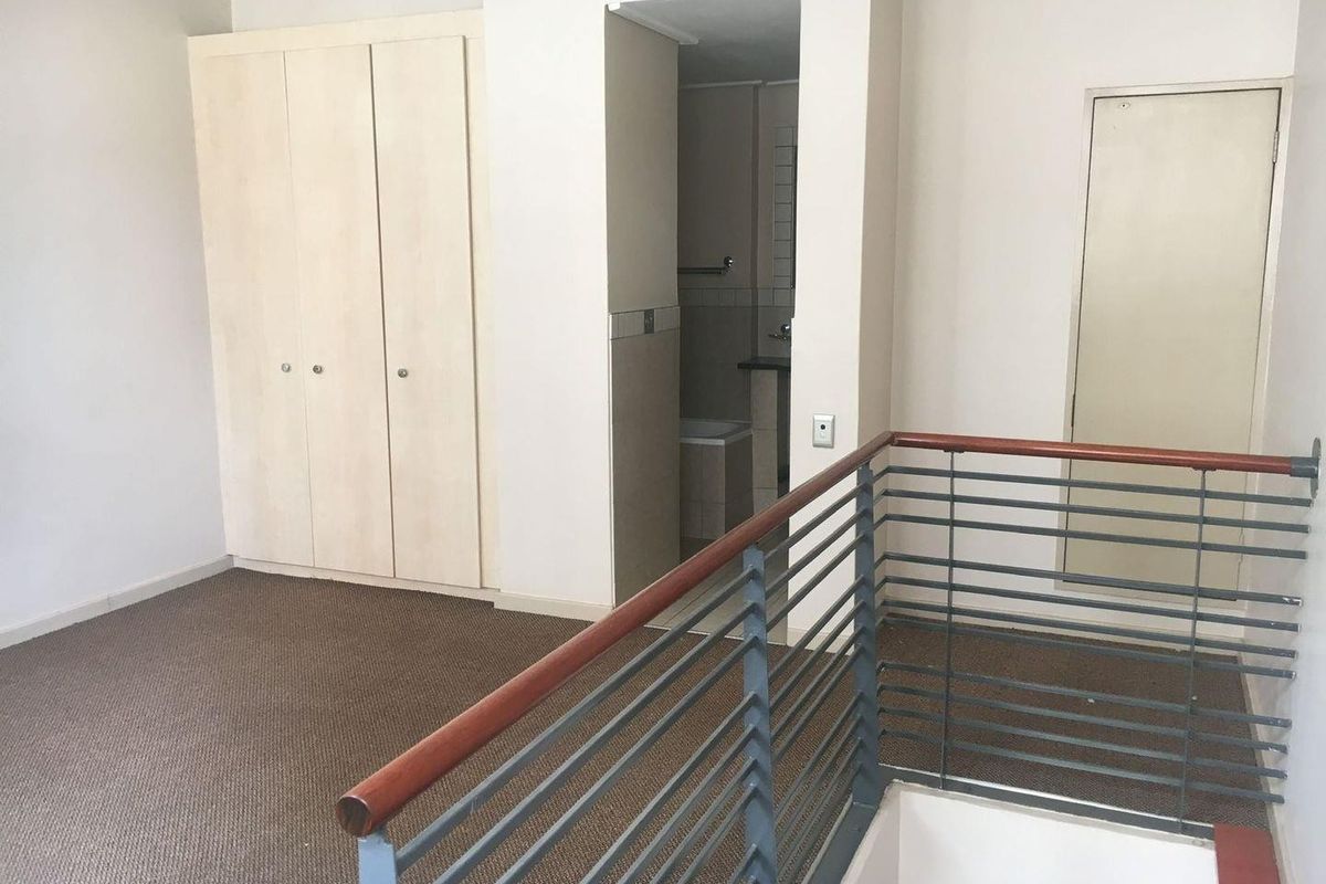 An empty bedroom featuring built-in cupboards, carpeted flooring, and a metal balustrade overlooking the staircase, with an adjacent en-suite bathroom.