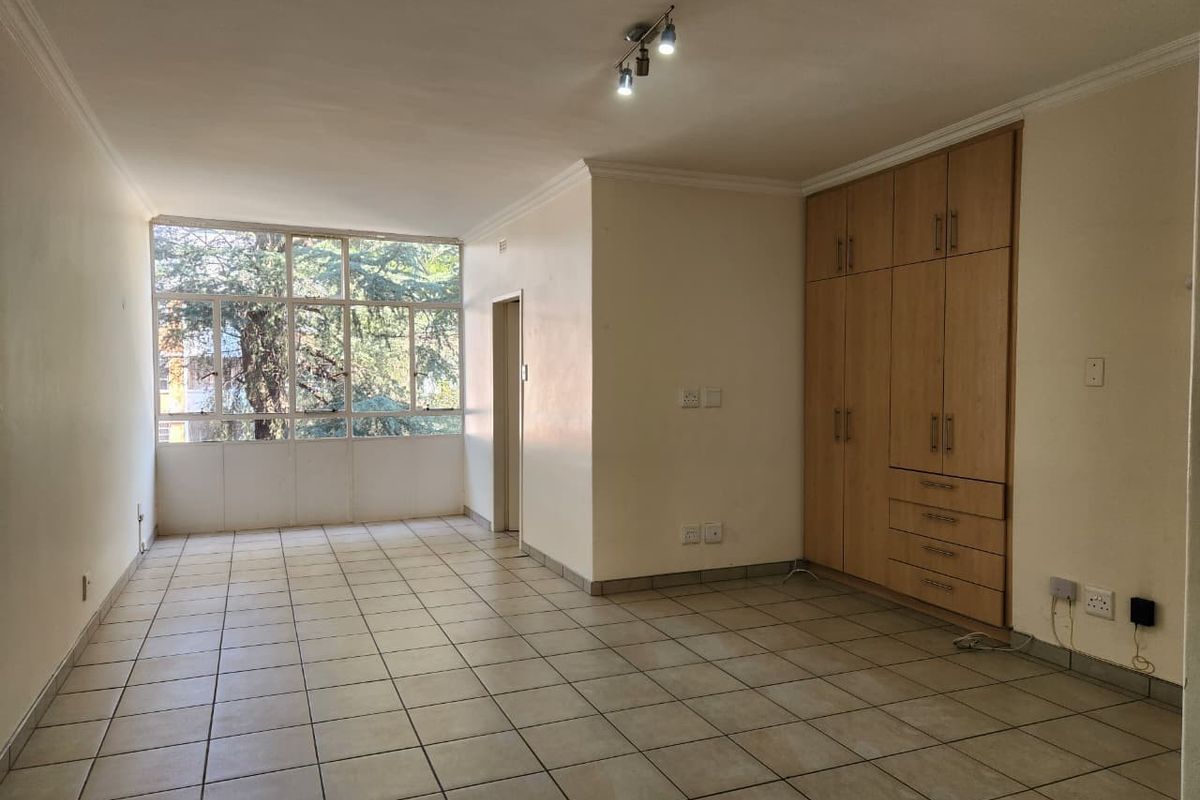 An empty living area featuring neutral tiled flooring, large windows providing natural light, and integrated built-in wooden cupboards for storage.