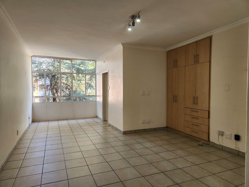 An empty living area featuring neutral tiled flooring, large windows providing natural light, and integrated built-in wooden cupboards for storage.