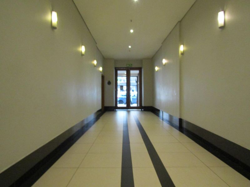 A clean, well-lit apartment hallway featuring tiled flooring with decorative border detailing and modern wall-mounted lighting fixtures leading to the main entrance.