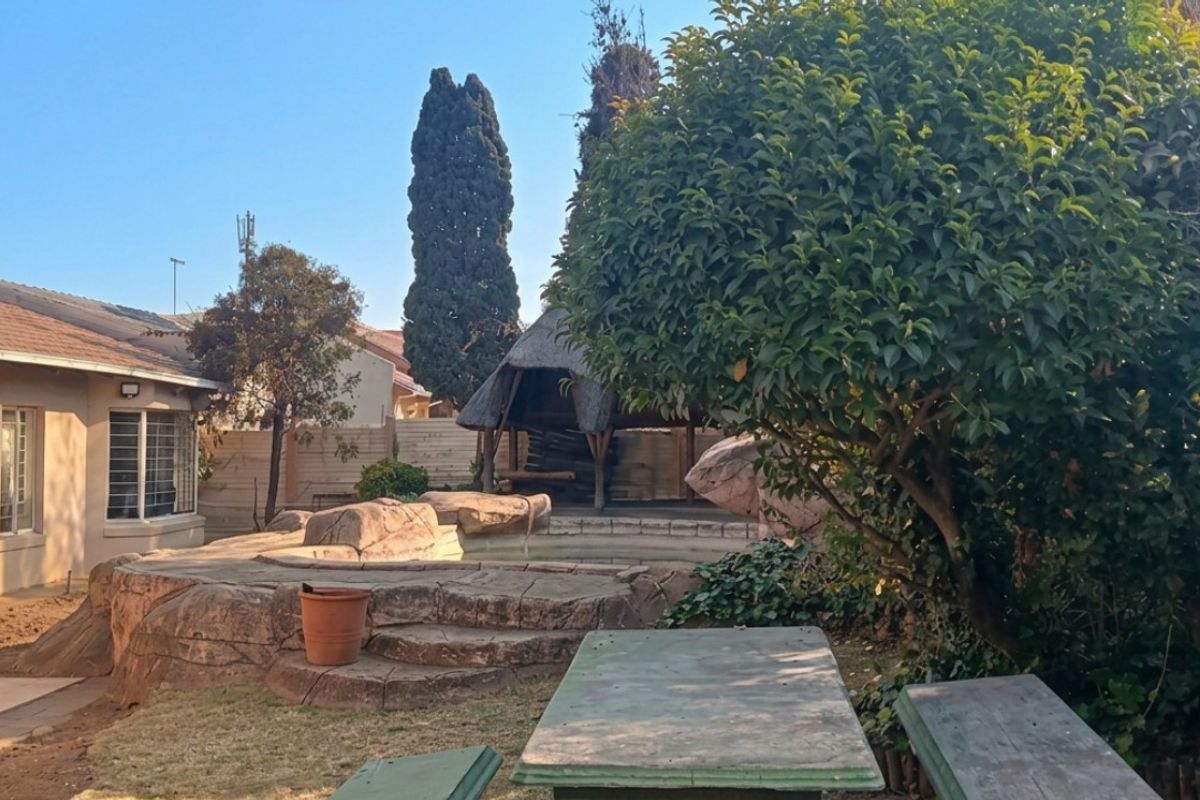 Rear garden area featuring a thatched lapa, decorative rock feature, and paved patio space adjacent to the main house structure.