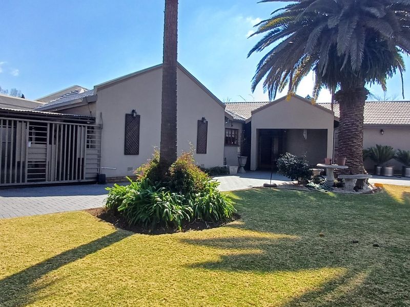 A well-maintained rear exterior view of the house featuring a paved driveway, landscaped garden, mature palm tree, and secure gated access.