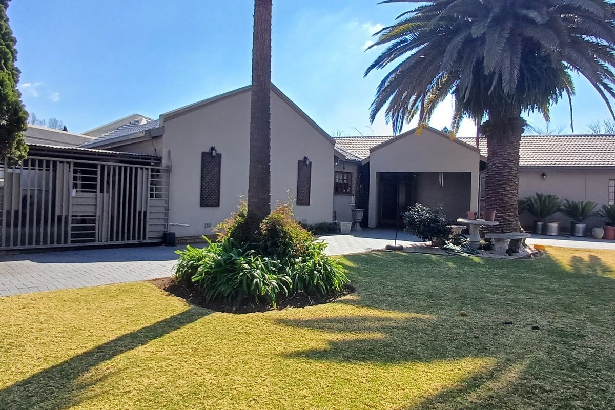 A well-maintained rear exterior view of the house featuring a paved driveway, landscaped garden, mature palm tree, and secure gated access.
