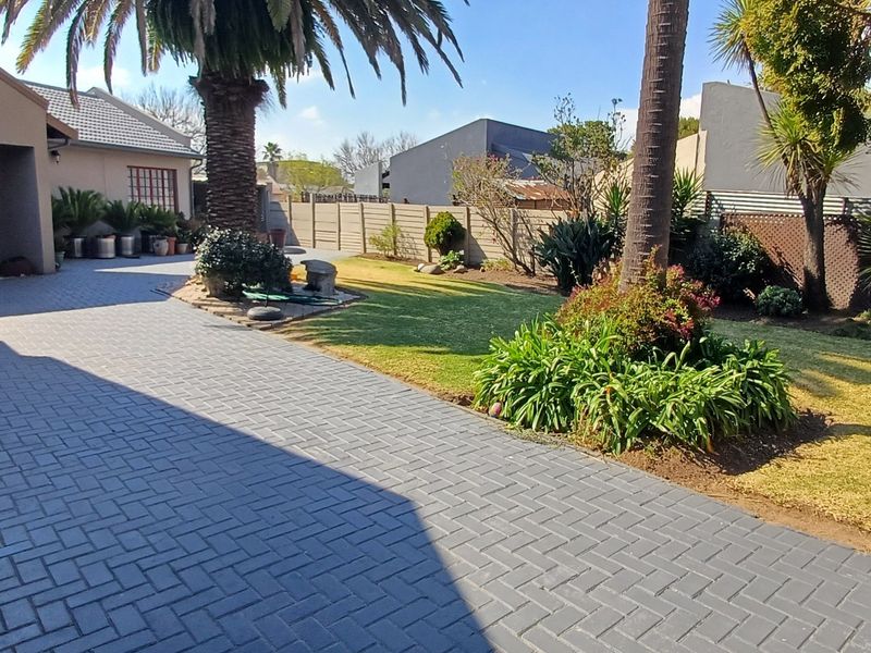 A well-maintained rear exterior featuring a paved driveway, covered patio area, and a landscaped garden with established trees and boundary walling.