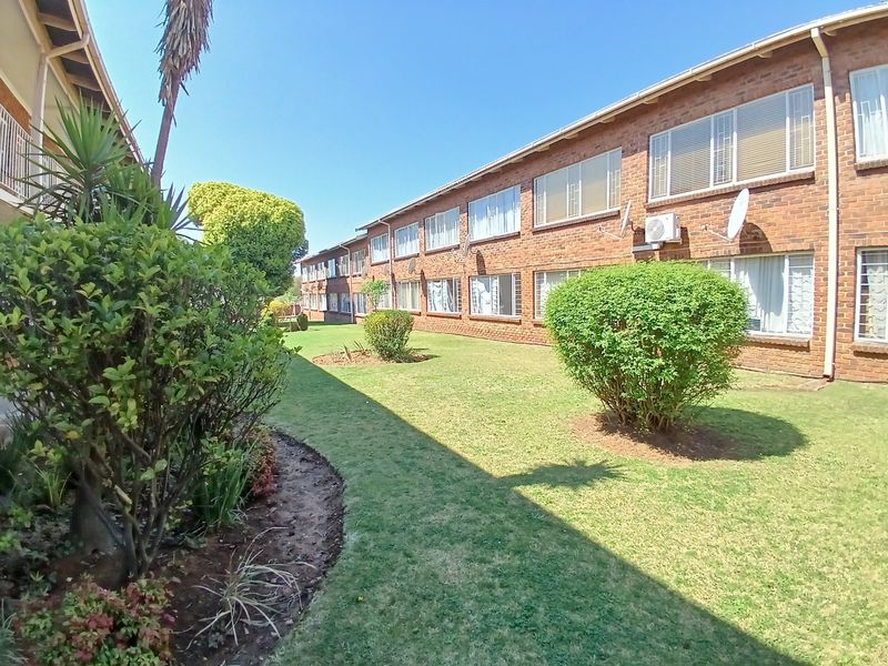 A view of the apartment complex exterior featuring a well-maintained communal garden, brick facade, and security-fitted windows.
