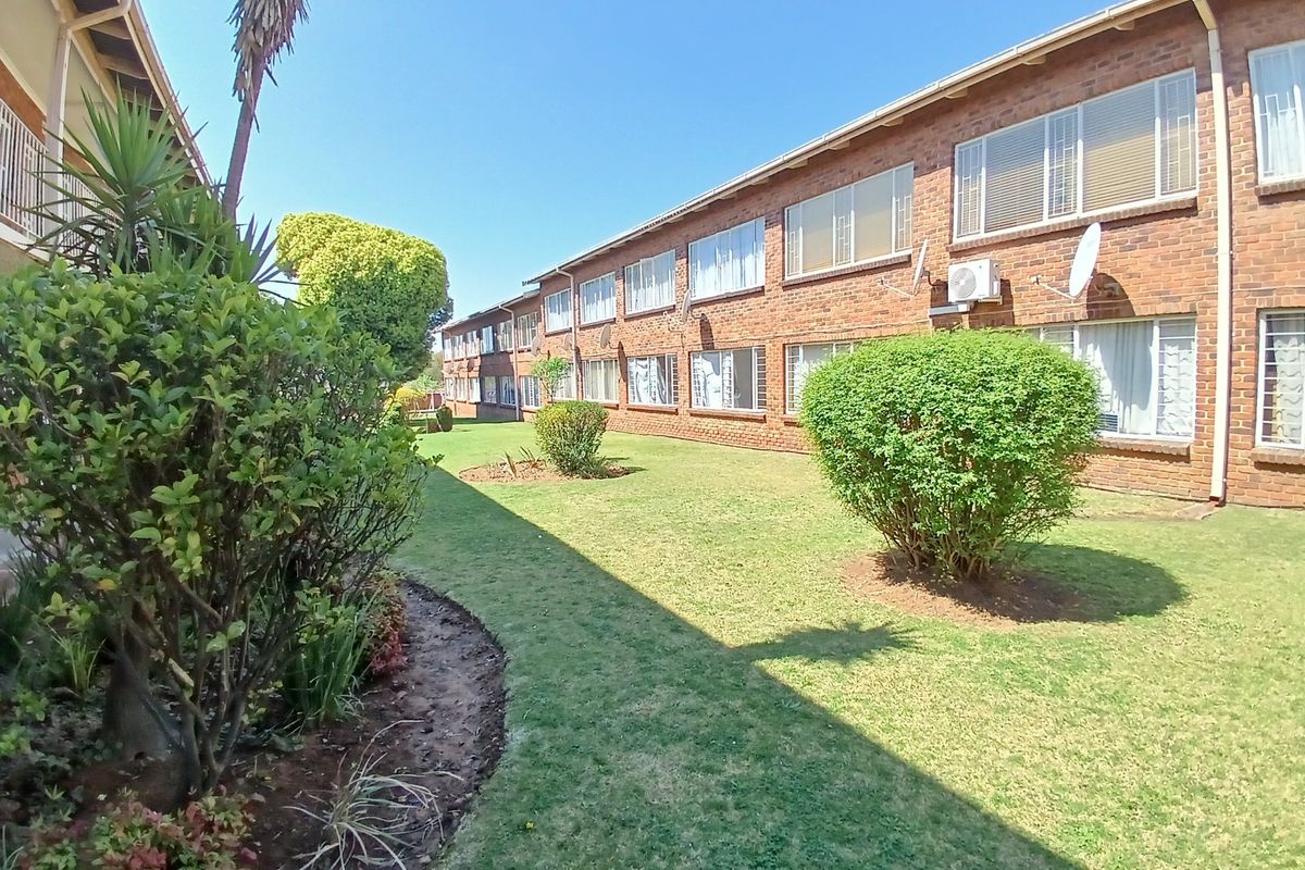 A view of the apartment complex exterior featuring a well-maintained communal garden, brick facade, and security-fitted windows.