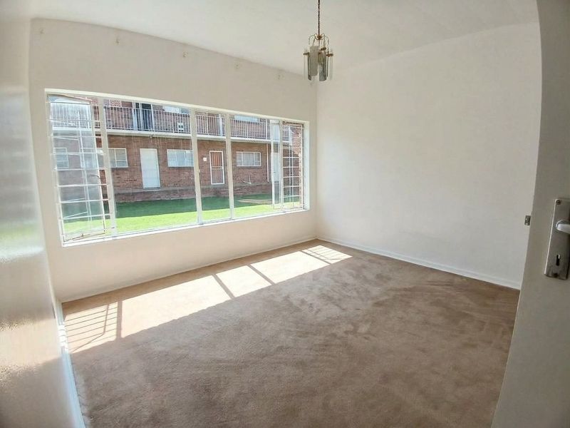 A bright, empty bedroom featuring large windows with security bars, neutral carpeting, and a central ceiling light fixture.